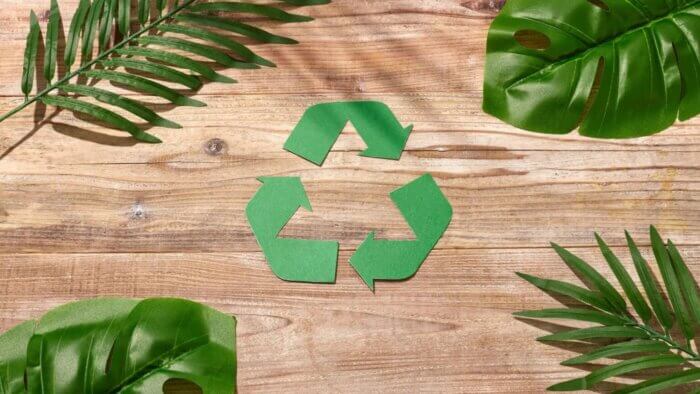 A recycle sign in front of a wooden background with green leaves next to it